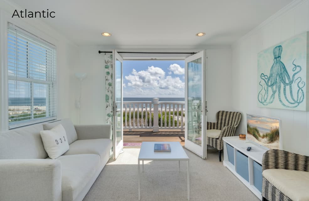A bright living area in a studio apartment featuring a light grey sofa with a decorative "931" pillow, a white coffee table, and striped armchairs. Large open French doors lead to a private balcony with a white railing, overlooking a sunny beach and the blue ocean under a clear sky.