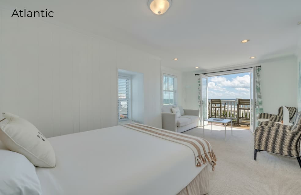 A bright suite viewed from the perspective of a bed with white linens and a striped throw blanket. In the background, open French doors lead to a balcony with a white railing, overlooking a sunny beach and a blue ocean under a partly cloudy sky.