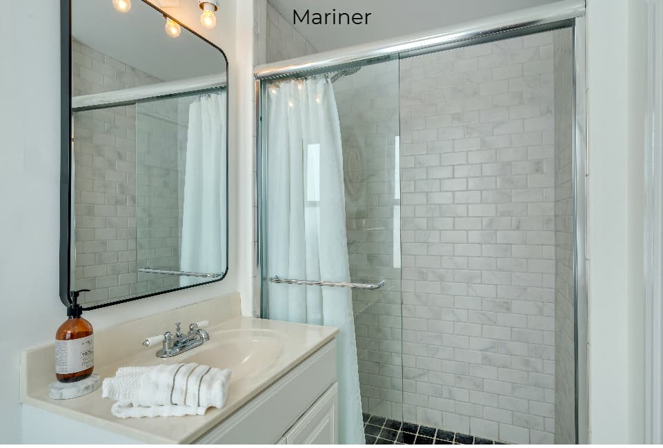 A bright, modern bathroom featuring a white vanity with a chrome faucet and a large rectangular mirror. To the left, a white toilet is positioned beneath a small white shelf and a circular woven wall decoration, while a frosted window provides soft natural light. On the right, a glass-enclosed walk-in shower is partially visible through an open white door.