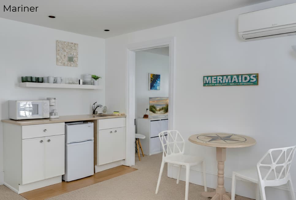 A brightly lit kitchenette area with white cabinets and light wood countertops featuring a microwave, coffee maker, and mini-fridge. To the right, a small round table with two white geometric chairs sits beneath a "MERMAIDS" wall sign, and an open doorway leads to a bedroom.