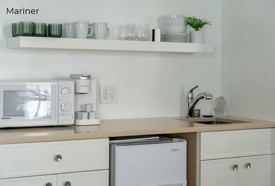 A white kitchenette with light wood countertops featuring a small sink, a coffee maker, and a white microwave. Above the counter, a white floating shelf holds glassware and small plants, while a compact white refrigerator is built into the lower cabinetry.
