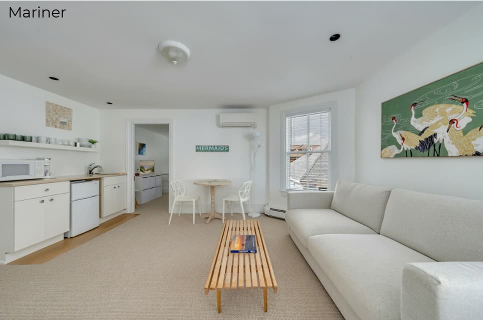A brightly lit, modern bathroom with white walls and a light tan tile floor. It features a white vanity with a chrome faucet and a large mirror, a white toilet, and a glass-enclosed walk-in shower with beige tiles. To the right, a large window with white blinds provides ample natural light.
