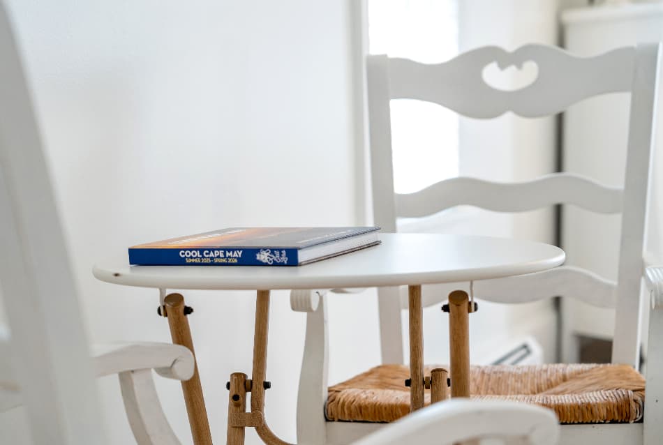 A close-up shot of a small, round white table with a thin wooden base. A blue book titled "Cape May" lies flat on the table's surface. In the background, a white wooden chair with a woven wicker seat is partially visible.