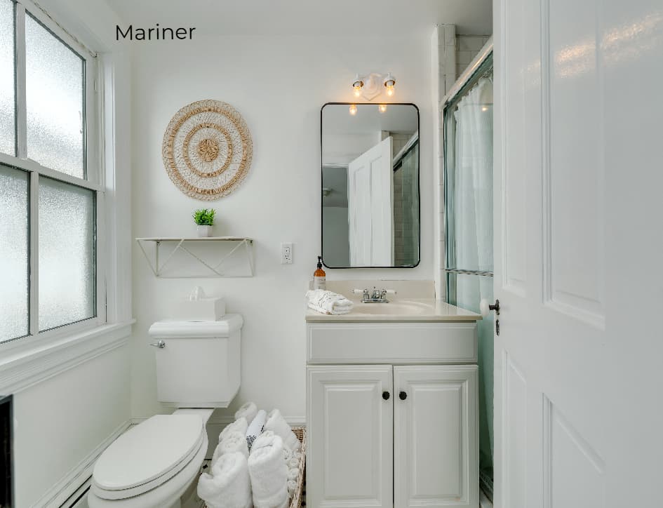 A bright, modern bathroom featuring a white vanity with a chrome faucet and a large rectangular mirror. To the left, a white toilet is positioned beneath a small white shelf and a circular woven wall decoration, while a frosted window provides soft natural light. On the right, a glass-enclosed walk-in shower is partially visible through an open white door.
