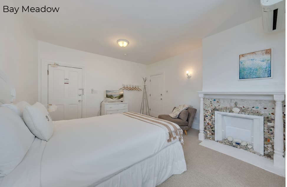 A white-walled bedroom featuring a white bed with a striped throw, a gray armchair, and a white mantle decorated with colorful mosaic tiles. In the background, a white door and a light-colored dresser are visible under a warm ceiling light.