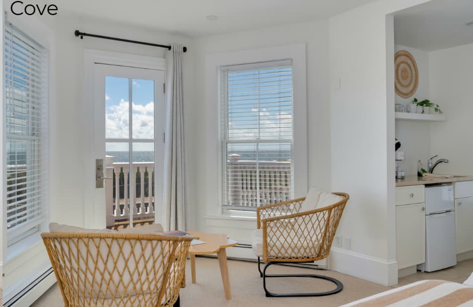 A view from inside the Cove suite looking out toward a white-railed balcony with two wooden chairs. In the foreground, two woven wicker chairs face a light-colored wooden coffee table positioned between two large windows and a glass door that reveal the ocean and a blue sky.