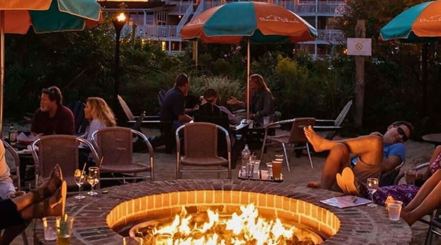 A cozy outdoor gathering around a fire pit with people enjoying drinks and conversation under patio umbrellas.