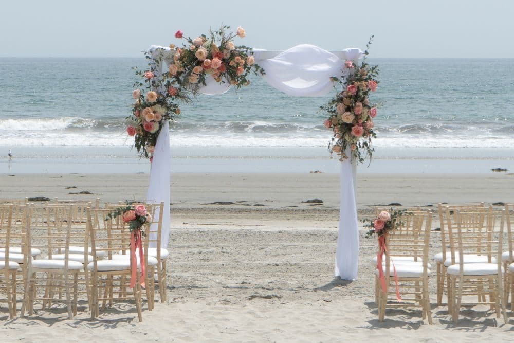 A wedding altar adorned with flowers sits on a sandy beach, facing the ocean with chairs arranged for guests.