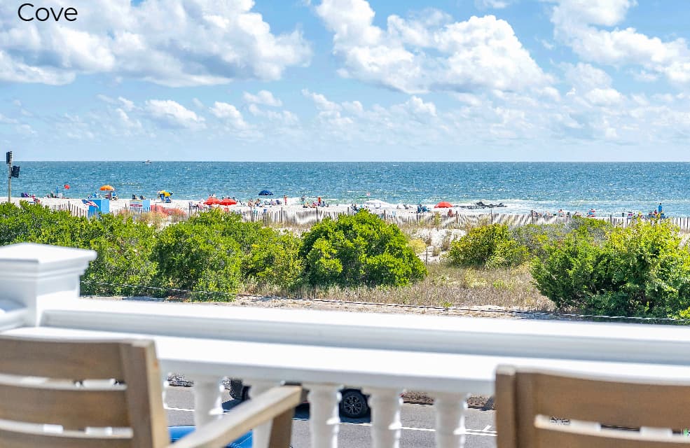 A wide-angle view from a wooden deck labeled "Cove," featuring a white railing with a built-in bar top and two wooden bar-style chairs. The balcony overlooks a green lawn and a sandy beach, with the blue ocean and a clear sky with scattered clouds in the background.
