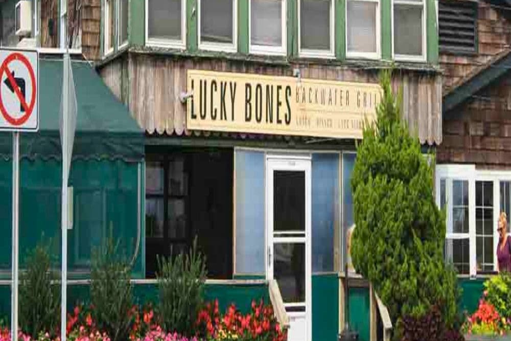 The exterior of Lucky Bones restaurant features a wooden facade and vibrant flowers, with a green canopy and a no U-turn sign nearby.