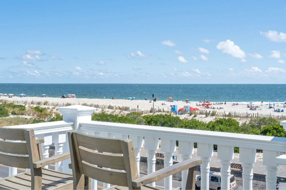 A view of a sandy beach and ocean from a deck with two wooden chairs.