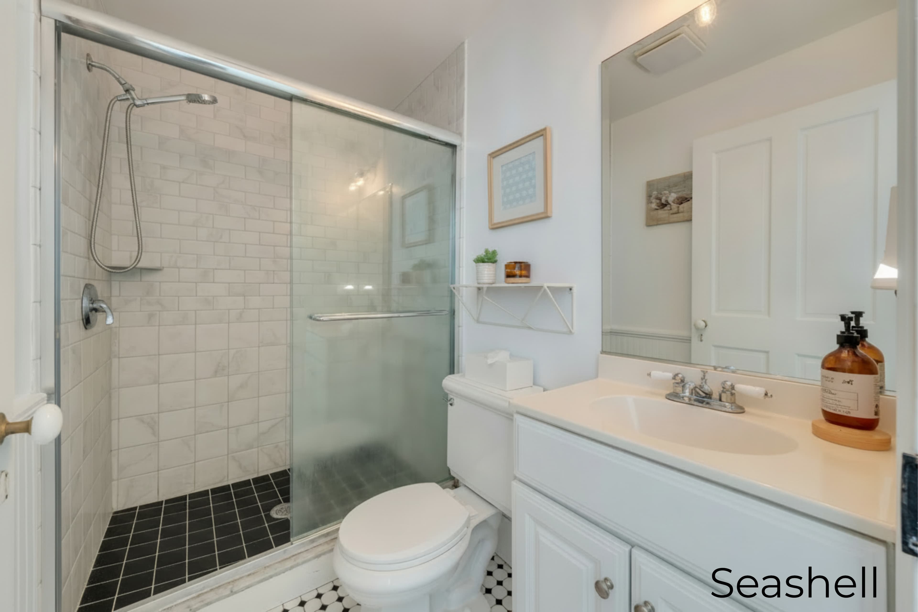 A brightly lit bathroom featuring a glass-enclosed walk-in shower with white subway tile walls and dark floor tiles. The room includes a white vanity with a large mirror, a white toilet, and minimalist decor on a small shelf.