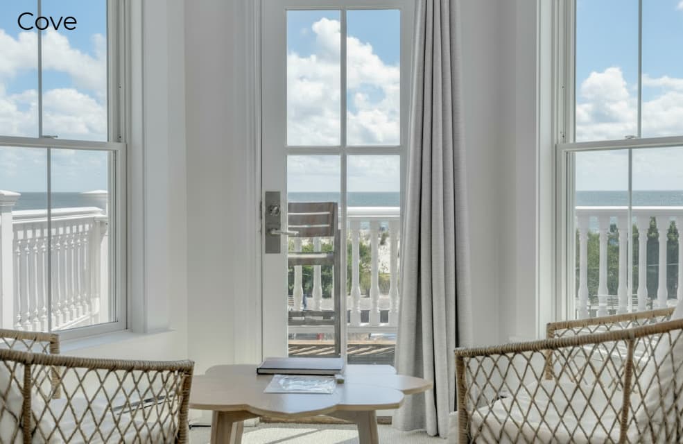 A view from inside the Cove suite looking out through large windows and a glass door onto a white-railed balcony with two wooden chairs. In the foreground, two woven wicker chairs face a small, light-colored wooden coffee table. The ocean and a bright blue sky with white clouds are visible in the background.