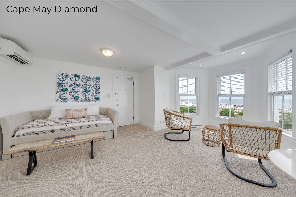 A spacious living area with white walls and light beige carpeting. The room features a neutral-toned daybed topped with a striped blanket and a small patterned pillow, set against a wall with a blue and white botanical art print. Two modern wicker armchairs are positioned near a large bay window that offers a view of the outdoors.
