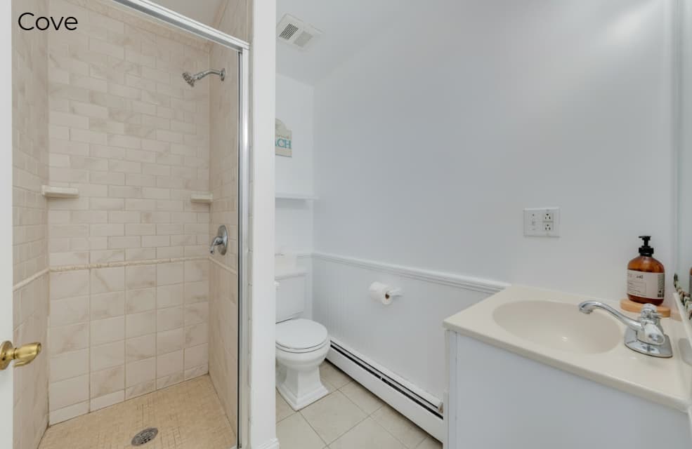 A simple white bathroom in the "Cove" suite featuring a walk-in shower with tan subway tiling and a glass door, a white toilet, and a small vanity with a cream-colored countertop.
