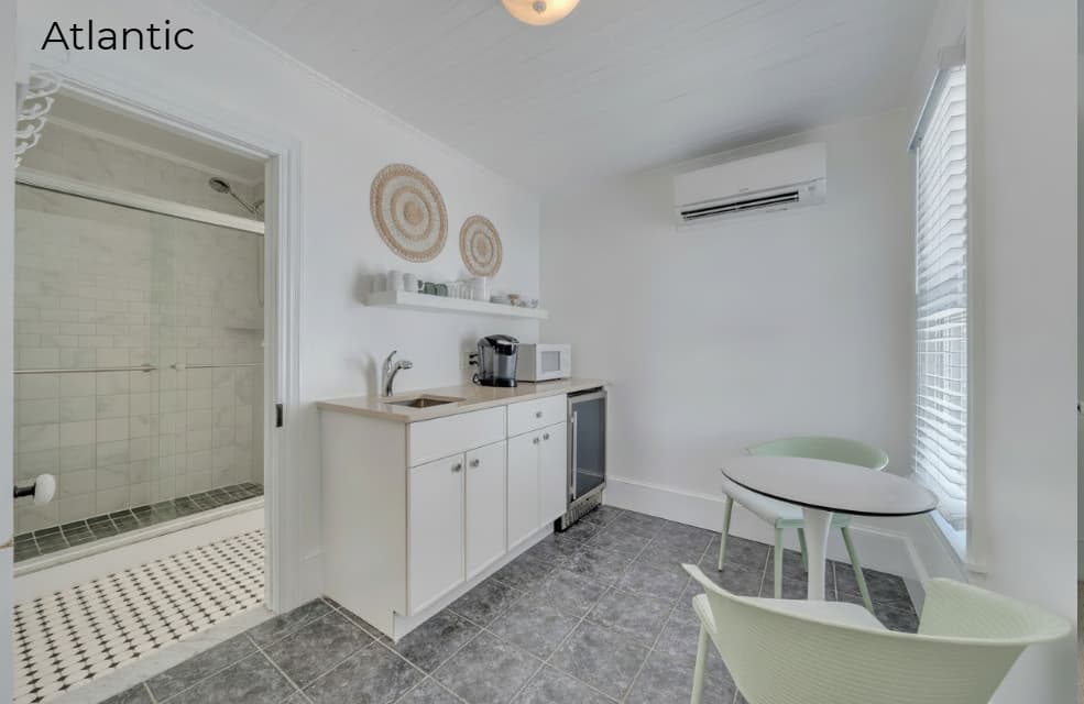 A brightly lit kitchenette featuring white cabinetry and light wood countertops, with a coffee maker and microwave. The space includes a small, round white dining table with two light green chairs, set against a window with white blinds and grey-tiled flooring. To the left, an open doorway reveals a modern bathroom with a glass-enclosed walk-in shower.