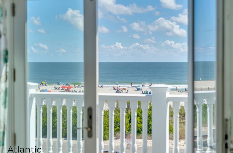 A view through open white balcony doors overlooking a sun-drenched beach and a calm blue ocean under a bright, partly cloudy sky. The white railing of the balcony is in the foreground, with green beach shrubs and colorful umbrellas on the sand visible in the distance.