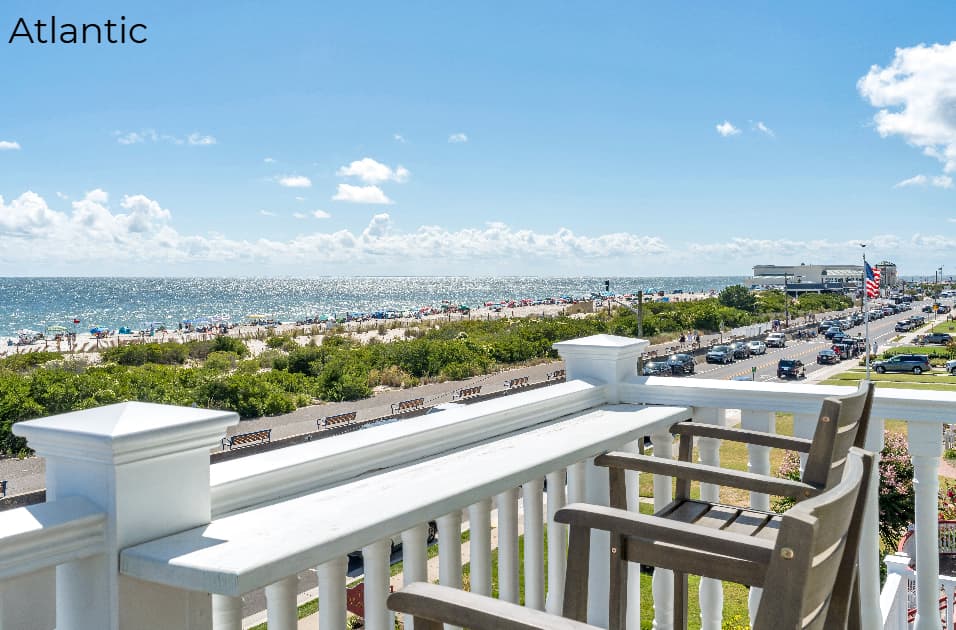 A wide-angle view from a second-story balcony featuring a built-in white bar top and two wooden high-top chairs. The balcony overlooks a coastal road lined with parked cars, a sandy beach with beachgoers and umbrellas, and the sparkling blue ocean under a bright, partly cloudy sky.