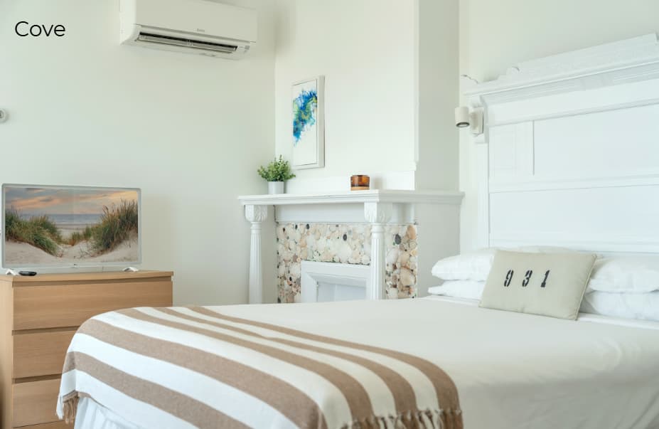 A minimalist, beach-themed bedroom labeled "Cove," featuring a white bed with a tan and white striped throw. The room includes a wooden dresser topped with a flat-screen TV displaying a beach scene and a white fireplace mantle decorated with multi-colored sea stones. A wall-mounted air conditioning unit is positioned on the white wall above the dresser.