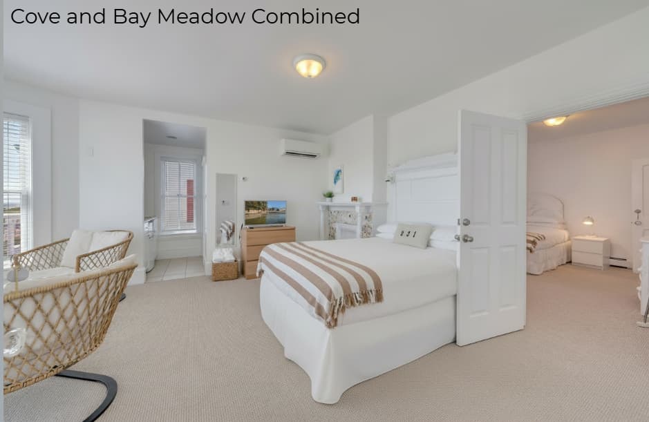 A primary white bed with a striped throw in the foreground and a second bed visible through an open white door in the background. The room includes woven wicker chairs on the left, a light-colored dresser with a TV, and a wall-mounted air conditioning unit.