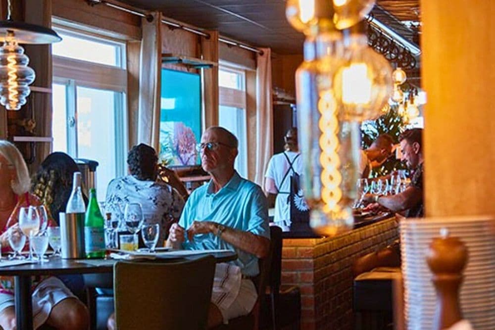 A man sits alone at a table in a warmly lit restaurant with other patrons in the background.